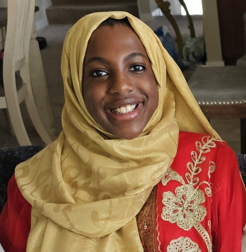 Photo of a woman with brown skin, brown eyes and black hair smiling wearing a gold fabric hijab and red patterned dress.