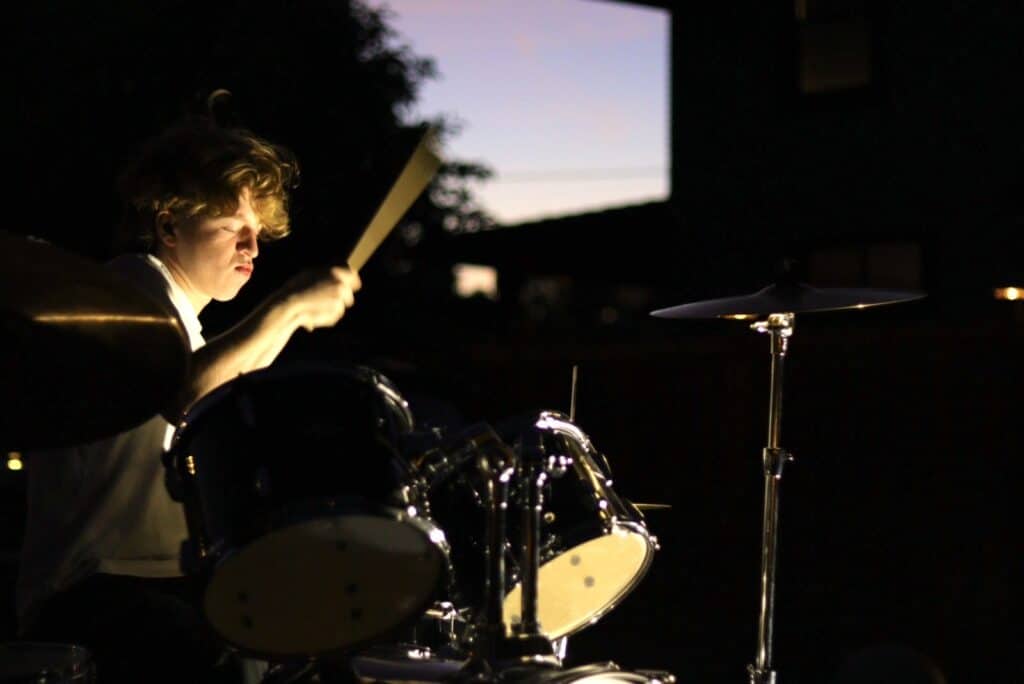 Photo of a young man holding a drum brush in the air. His face is illuminated and shows his light hair and skin. There is a drum under the drum brush with a light underside that shows. There is light purple sky showing in the background behind a dark outline of an evergreen tree.