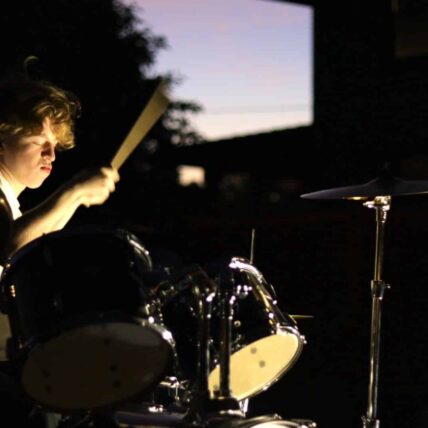 kai (1) Photo of a young man holding a drum brush in the air. His face is illuminated and shows his light hair and skin. There is a drum under the drum brush with a light underside that shows. There is light purple sky showing in the background behind a dark outline of an evergreen tree.