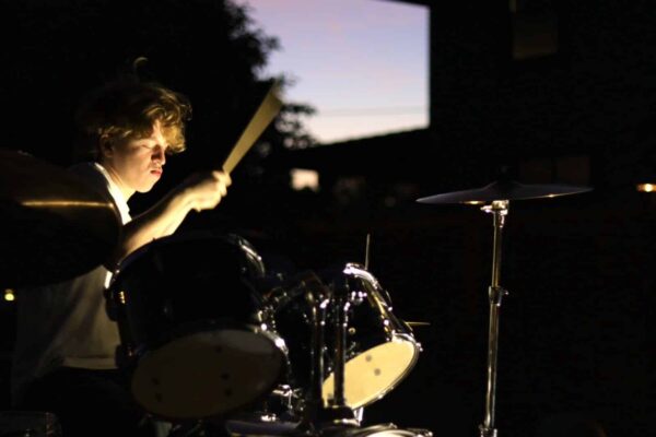 Photo of a young man holding a drum brush in the air. His face is illuminated and shows his light hair and skin. There is a drum under the drum brush with a light underside that shows. There is light purple sky showing in the background behind a dark outline of an evergreen tree.