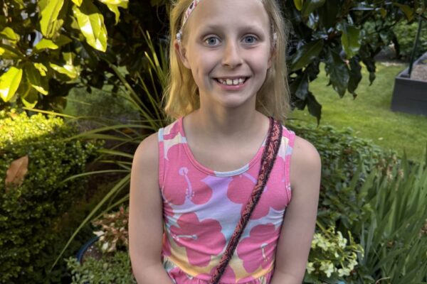 Photo of a school aged girl with shoulder length blonde hair and blue eyes. She is smiling and wearing a lilac sleeveless dress with a large pink flower print. There are large green leafy plants in the background.