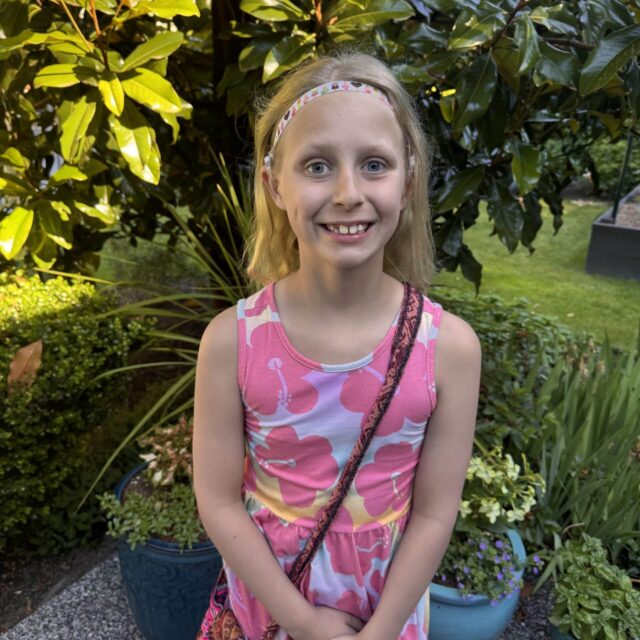 marna Photo of a school aged girl with shoulder length blonde hair and blue eyes. She is smiling and wearing a lilac sleeveless dress with a large pink flower print. There are large green leafy plants in the background.