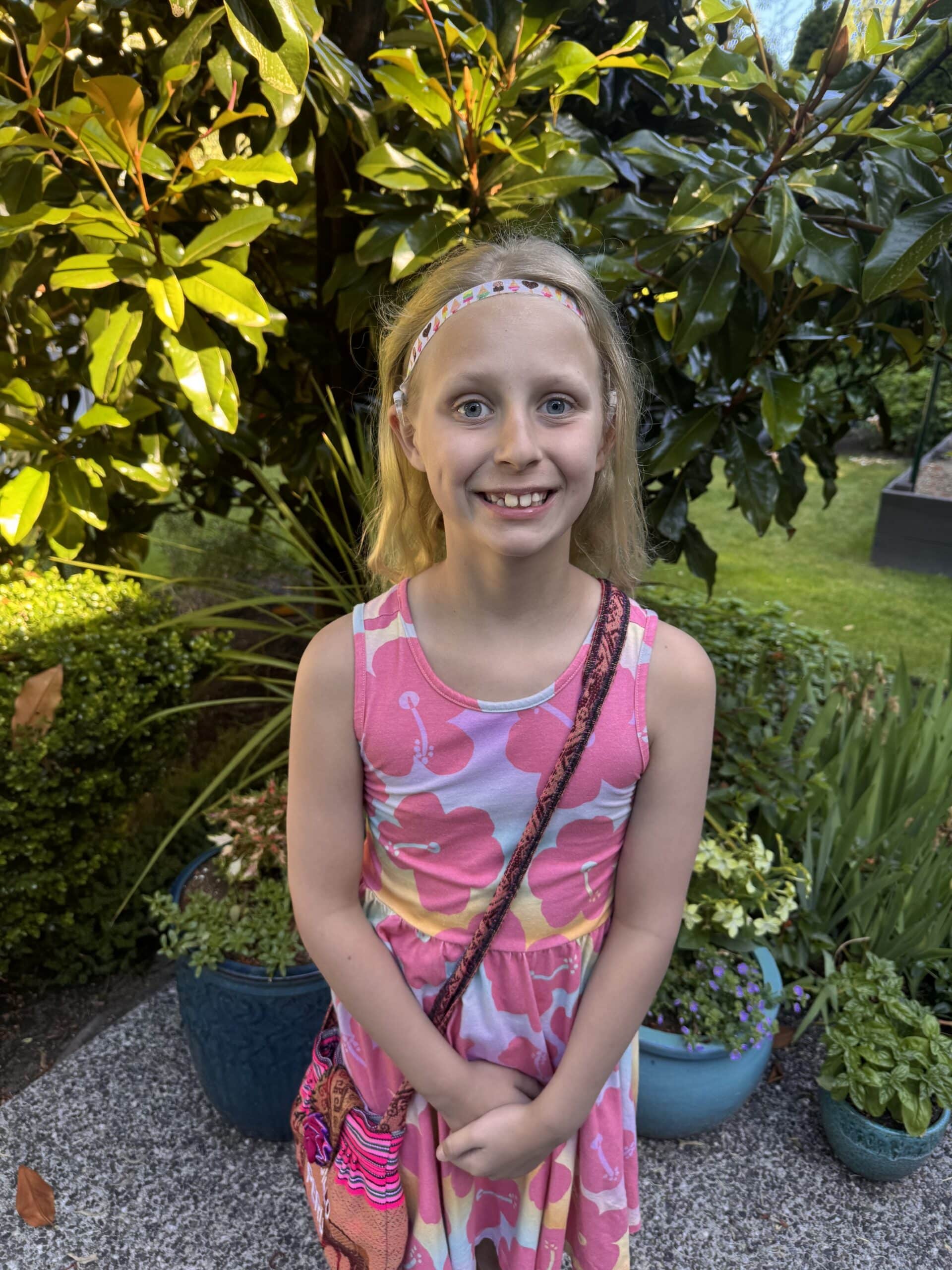 Photo of a school aged girl with shoulder length blonde hair and blue eyes. She is smiling and wearing a lilac sleeveless dress with a large pink flower print. There are large green leafy plants in the background.